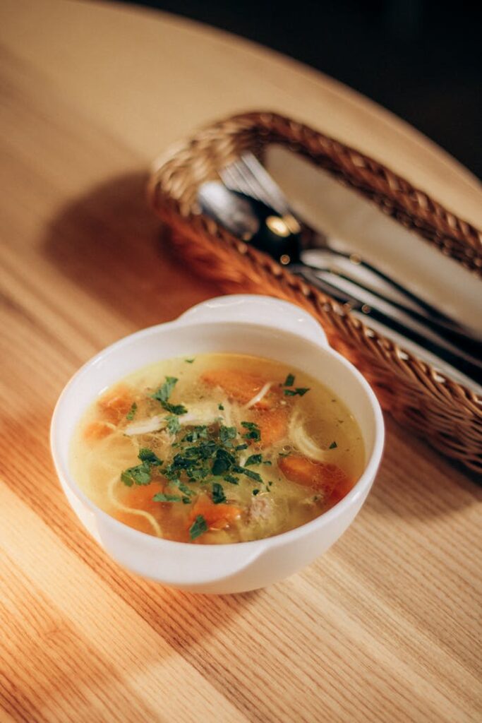 Delicious homemade chicken soup served hot in a white bowl on a wooden table with cutlery basket.